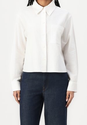 Femme portant une chemise blanche unie à boutons avec une poche et un jean bleu foncé, debout devant un fond blanc.