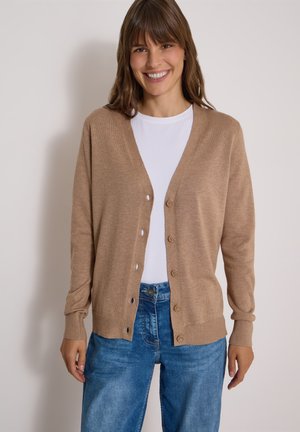 Vrouw glimlacht en draagt een bruine gebuttonte cardigan over een wit overhemd en een blauwe spijkerbroek, staand tegen een effen lichte achtergrond.