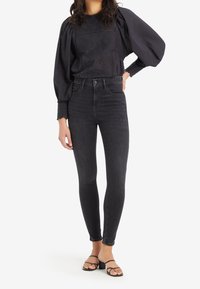 Blusa negra de manga abullonada con bordado floral, combinada con unos pantalones skinny negros de cintura alta y sandalias negras de tiras. Textura suave en toda la prenda.