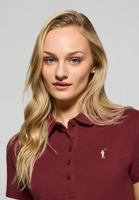 Bordowa koszulka polo z krótkim rękawem, z trzema guzikami z przodu i haftowanym logo na lewej piersi. Materiał bawełniany, strukturalne wykończenie.