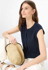 Donna con lunghi capelli castani che indossa un gilet lavorato a maglia senza maniche blu navy e pantaloni bianchi, che tiene un cappello beige intrecciato con bordo nero.