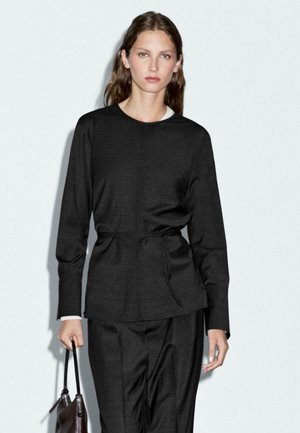 Black textured blazer with a tie waist, paired with matching trousers. Underneath, a white shirt collar is visible. Model holds a dark handbag.