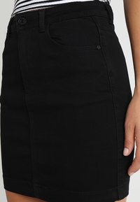 Gros plan sur une personne portant une jupe en denim noir et un haut rayé, mettant en évidence la poche avant et les détails des boutons de la jupe.