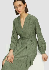 Abito lungo verde realizzato in tessuto ad occhiello, con orlo spacco, chiusura con bottoni e vita con cintura. Presenta un design leggero e strutturato.