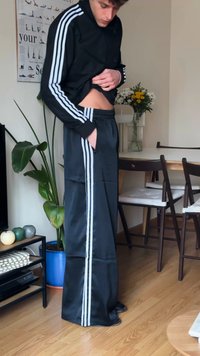 Svart treningsjakke med hvite striper, matchende svarte bukser med sidestiper, brukt i en avslappet innendørs setting med planter.