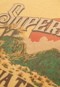 Tissu jaune avec un motif graphique vintage représentant des montagnes vertes, des cactus et un coucher de soleil aux tons rouges et orange. Texte : "Super."