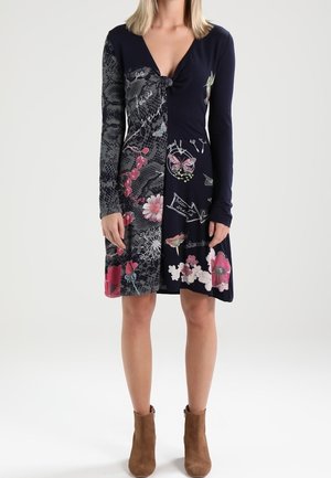 Frau trägt ein marineblau-schwarzes Patchworkkleid mit Blumen- und Schmetterlingsmotiven, kombiniert mit braunen Stiefeletten, vor weißem Hintergrund stehend.