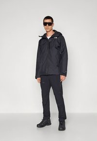 Crna vodootporna jakna s kapuljačom, nošena preko odgovarajućih hlača. Tkanina izgleda glatko, s minimalnim detaljima i džepom na patentni zatvarač na jakni.