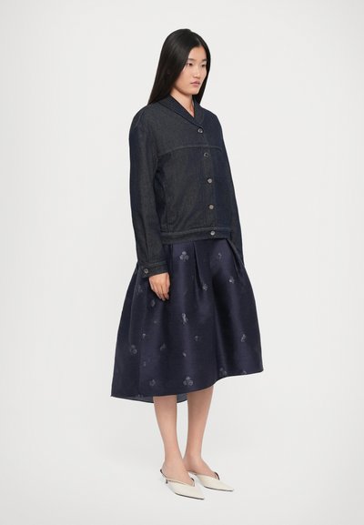 Woman wearing a dark denim jacket over a navy midi skirt with subtle floral patterns and white pointed-toe mule heels, standing on a plain background.