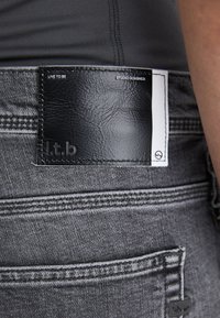 Sive traperice s crnim sintetičkim kožnim detaljem. Tekst: "ŽIVI DA BI BIO" i "STUDIO DIZAJNIRANO." Imaju detalje šivanja i oznaku.