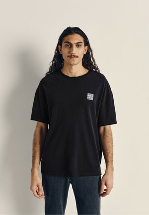 T-shirt en coton noir à manches courtes, col rond, avec un petit patch logo blanc sur la poitrine ; porté avec un jean foncé.