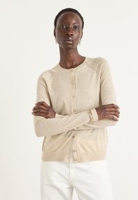 Beige stickad cardigan med rund halsringning, knäppning framtill och långa ärmar. Slät textur, ribbade detaljer och en avslappnad passform.