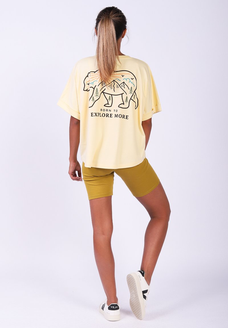 Gelbes, Übergröße-T-Shirt mit Bärenmotiv, Bäumen und Bergen auf dem Rücken. Kombiniert mit senfgelben Shorts und weißen Sneakers.