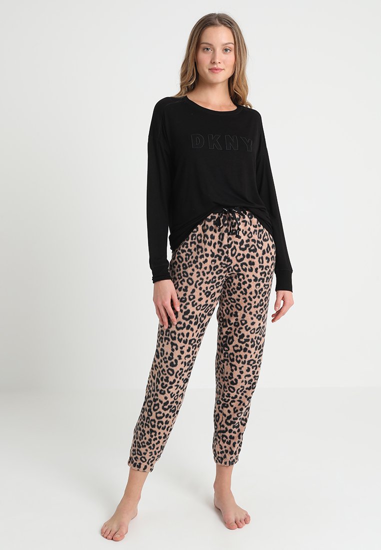 Młoda kobieta stojąca boso, ubrana w czarną bluzę DKNY z długim rękawem i beżowe piżamowe spodnie w wzór lamparta na białym tle.