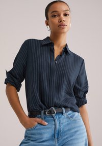 Námornícka modrá košeľa na gombíky s bielymi zvislými pruhmi, s vyhrnutými rukávmi a voľným strihom, nosená s svetlomodrými riflemi s vysokým pásom a čiernym opaskom.