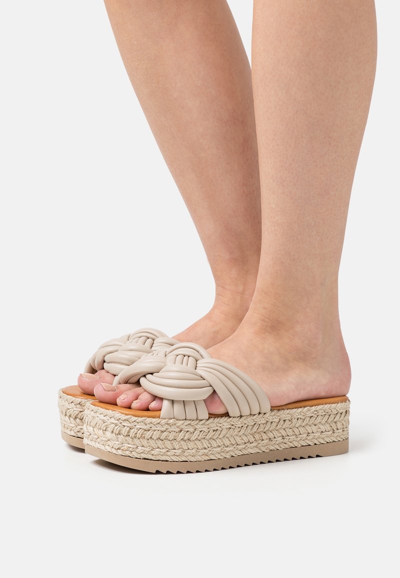 Pieds portant des sandales compensées beiges avec des semelles en corde tressée et des brides en cuir nouées et rembourrées sur un fond uni.