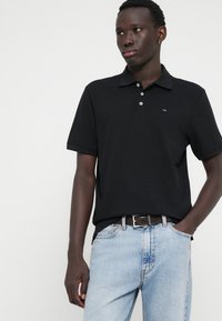 Must musta särk klassikalise kraega, millel on kolm nuppu ja väike logo rinna peal, kombineeritud helesiniste denim teksadega.
