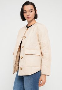 Πανωφόρι από καπιτονέ, ανοιχτού μπεζ χρώματος με στρογγυλή λαιμόκοψη, εμπρόσθιες τσέπες και κουμπιά σε χρυσό τόνο, με casual, άνετη εφαρμογή και υφή υφάσματος.