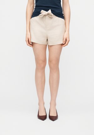 Kvinde iført beige højtaljede shorts med sløjfe foran, navyblå top og mørkebrune spidse hæle, stående mod en ensfarvet lys baggrund.