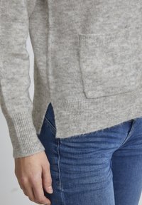 Pull en tricot gris clair avec poche avant, poignets côtelés et fentes sur les côtés, associé à un jean ajusté bleu foncé.