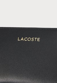 Billetera de cuero negro con superficie texturizada. Marca "LACOSTE" en dorado destacada en la parte frontal. Diseño elegante y minimalista.
