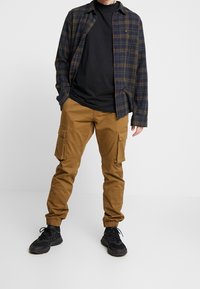 Geruite flanellen overhemd in marineblauw en groen, zwart t-shirt eronder, en tan cargo broek met elastische boorden; zwarte sneakers maken de outfit compleet.