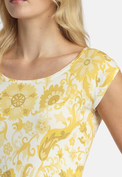 Nahaufnahme einer Frau in einem weißen Oberteil mit gelben Blumen- und Rankenmustern, die den Ausschnitt, die Schulter und einen Teil des Arms zeigt.