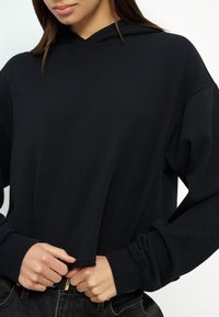 Femme ajustant l'ourlet d'un sweat à capuche noir surdimensionné, portant un jean foncé, sur un fond uni.