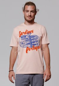 Hellrosa Baumwoll-T-Shirt mit einem blauen Sardinenbüchse-Motiv und orangefarbener Schrift mit der Aufschrift "Sardinen Portugal", kurzen Ärmeln und rundem Halsausschnitt.