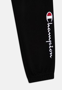 Juodi sportiniai kelniai, pagaminti iš minkštos medžiagos, su balta išsiuvinėta "Champion" logotipu ir raudonu bei baltu akcentu logotipe.