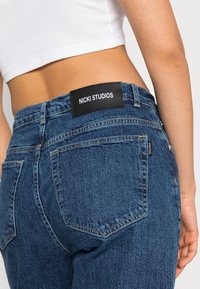 Jeans de ganga azuis com um corte de altura média, apresentando um rótulo de pele preta na cintura e costura dourada em contraste. Duas bolsas traseiras visíveis.