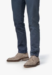Bottines en feutre marron avec un bout arrondi, des détails de lacets et une semelle contrastante, portées avec un pantalon ajusté bleu foncé.