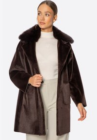 Abrigo de piel sintética marrón con un cuello amplio, corte oversized, dos bolsillos frontales y una textura suave, combinado con una parte superior y pantalones de color claro.