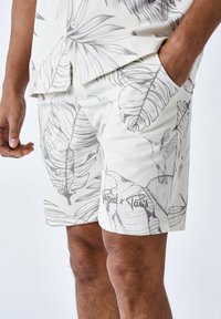 Pantalones cortos blancos con un estampado de hojas tropicales grises y colibríes. Están hechos de una tela ligera, con bolsillos laterales y un ajuste casual.