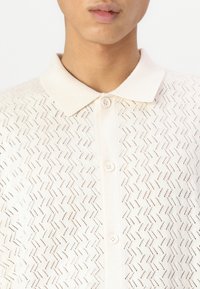 Cardigã de malha em bege claro com um padrão texturizado em zigzag. Apresenta um fecho de botão e um colarinho clássico. Tecido macio e respirável.