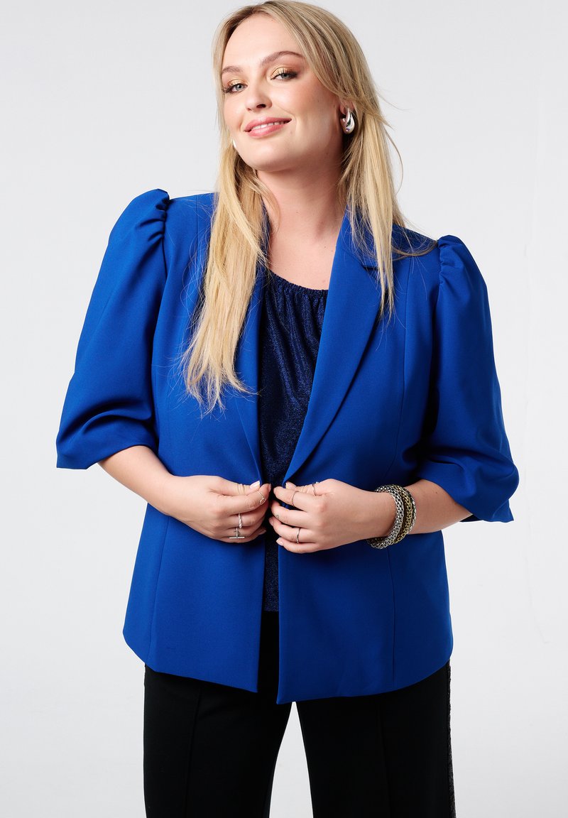Blauwe blazer met opstaande mouwen, gemaakt van een gladde stof. De blazer is open, waardoor een donkere top eronder zichtbaar is. Zwarte broek en getextureerde accessoires zijn ook zichtbaar.
