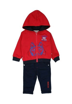 Haut à capuche zippé rouge avec un graphique de loup en bleu et un pantalon marine avec les poignets retroussés. Le matériau est doux, avec des accents noirs sur le haut à capuche.