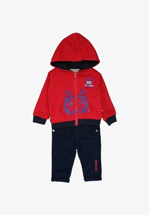 Haut à capuche zippé rouge avec un graphique de loup en bleu et un pantalon marine avec les poignets retroussés. Le matériau est doux, avec des accents noirs sur le haut à capuche.