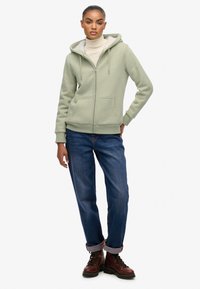 Lichtgroene zip-up hoodie met een ruime capuchon, zachte binnenvoering en voorzakken. Gepaard met opgerolde denim jeans en bruine laarzen.