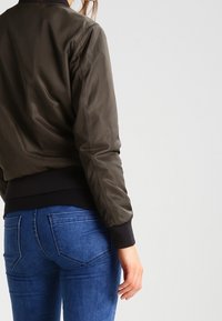 Olivgrön bomberjacka med en slät textur, ribbade cuffs och fåll. Tillsammans med blå denimbyxor som visar en åtsittande silhuett.