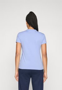 O femeie cu păr lung împletit poartă un tricou cu mânecă scurtă de culoare albastră deschisă și pantaloni de culoare albastră închisă, stând cu spatele îndreptată către un fundal simplu.