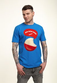 Blå t-shirt med en stor grafik av en Babybel-ost rundel med ett bett taget. Texten "OM NOM NOM" är i en röd pratbubbla.