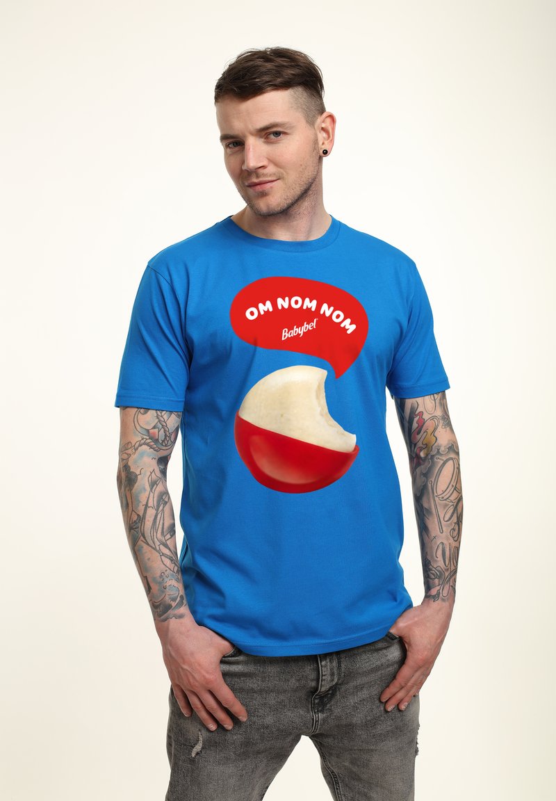 Blå t-shirt med en stor grafik av en Babybel-ost rundel med ett bett taget. Texten "OM NOM NOM" är i en röd pratbubbla.
