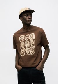 Mann trägt beige Kappe, braunes T-Shirt mit neun stilisierten alten Maskenzeichnungen und dem Text "the new originals", Hände in den schwarzen Hosentaschen.