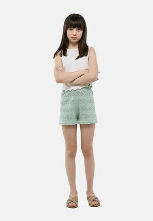Haut sans manches blanc avec des bords en losange et short en tricot vert menthe avec un motif en zigzag. Porté avec des sandales tressées. Debout les bras croisés.
