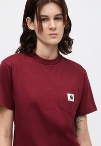 T-shirt in cotone borgogna con scollatura rotonda e maniche corte. Presenta una tasca sul lato sinistro del petto con un'etichetta del logo. Texture liscia e vestibilità comoda.
