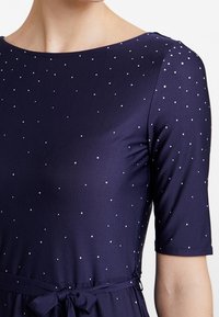 Námořnické šaty s krátkými rukávy, zaobleným výstřihem a drobnými stříbrnými tečkami jako ozdobami. Obsahují pásek v pase pro stažení.