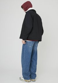 Veste noire avec une capuche en fourrure blanche au-dessus d'un sweatshirt bordeaux, associée à un jean bleu ample et des baskets de couleur claire.