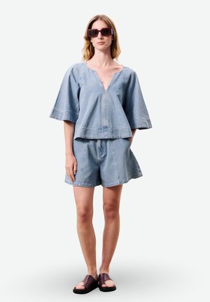 Femme portant un haut à manches courtes texturé bleu clair et un short assorti, lunettes de soleil foncées et sandales slides marron foncé, vue de face.