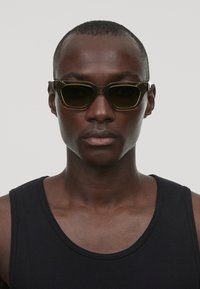 Lunettes de soleil à monture noire avec verres sombres, présentant une forme géométrique unique et un design angulaire. Portées avec un débardeur noir.